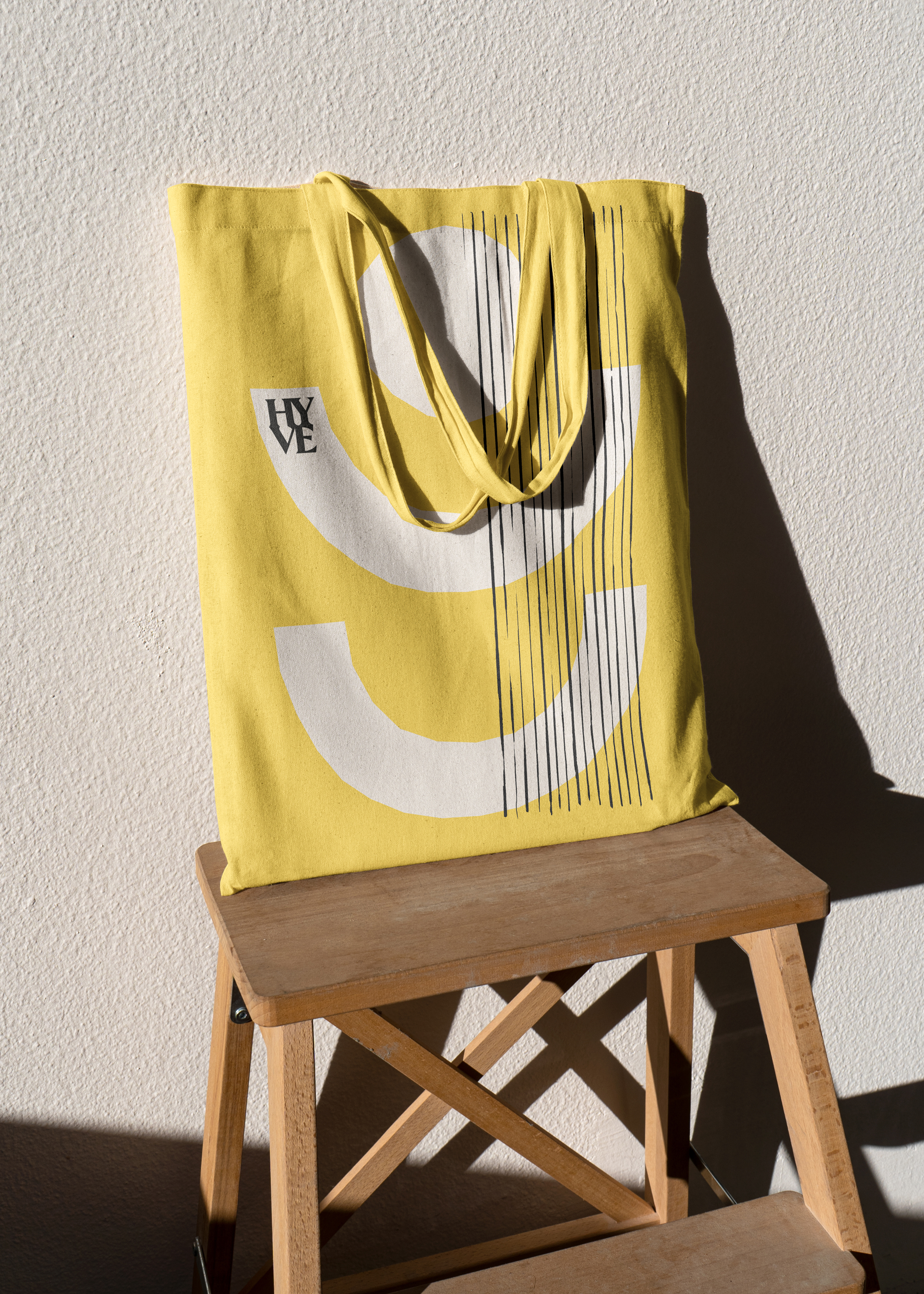 Black and white abstract brand shapes, printed onto a yellow tote bag, which is laying on a wooden stool.