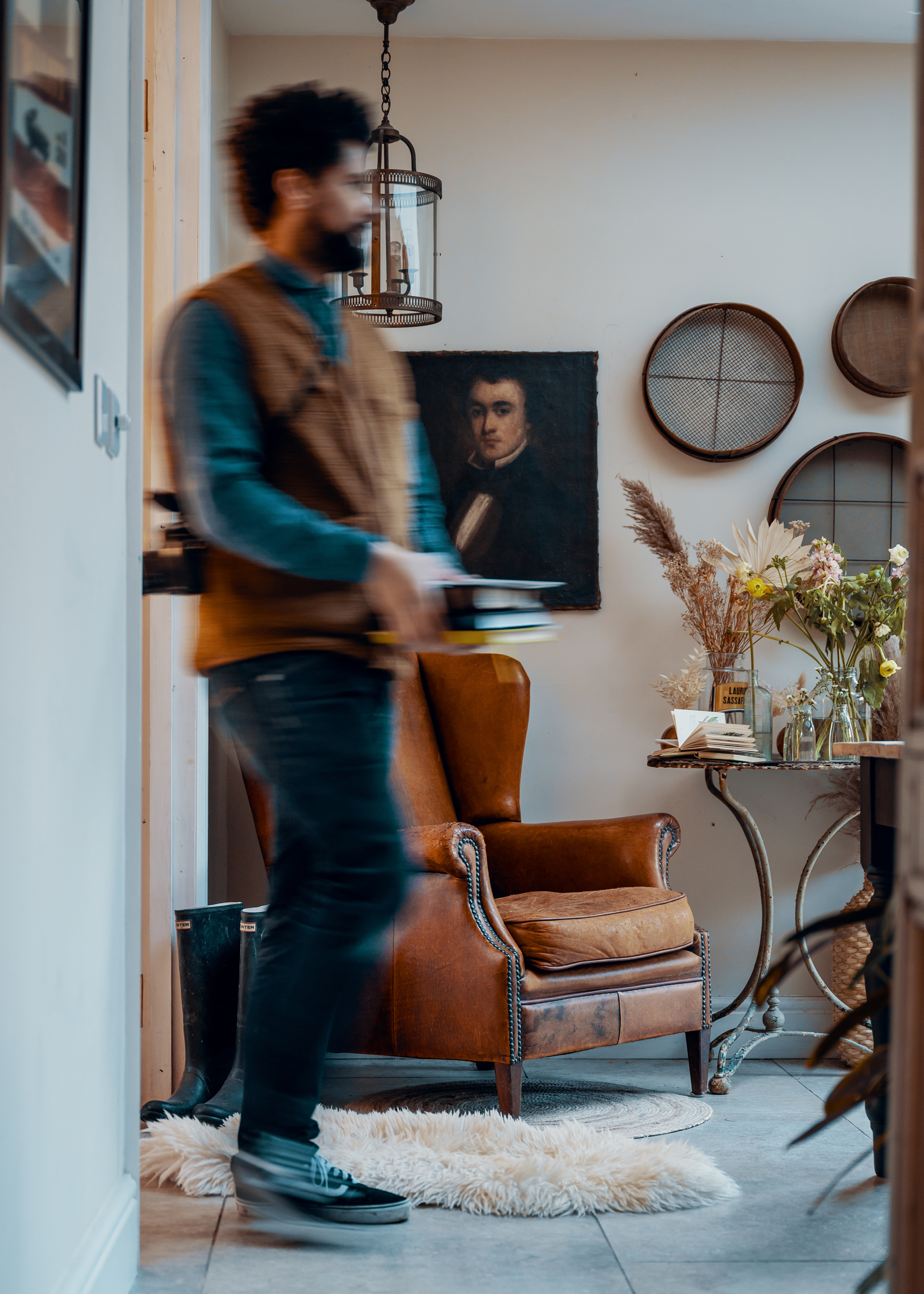 A well used brown leather chair in the corner of a room in a country house, with a pair of wellington boots sitting next to it. A man is walking in front of the chair, blurred by his motion.