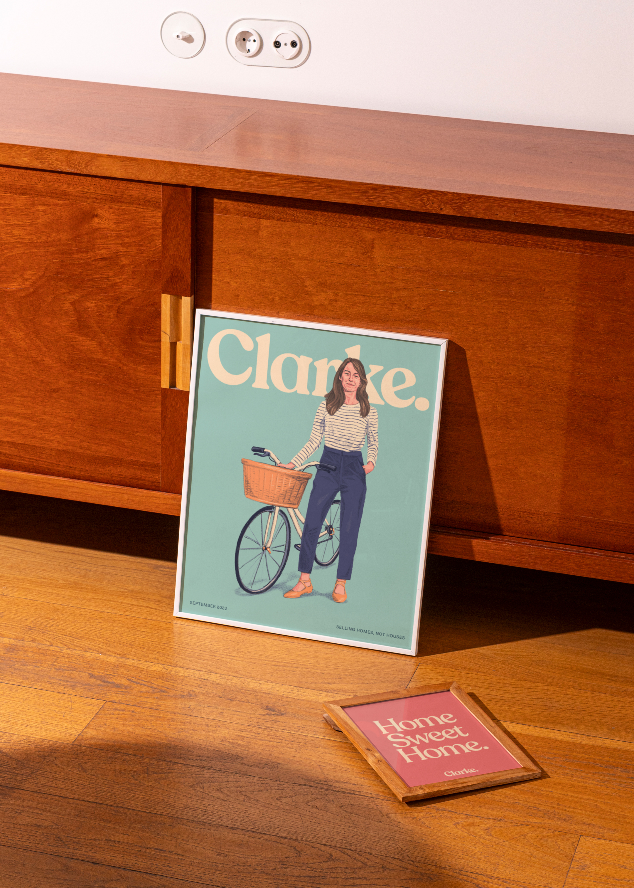 Two framed posters. One showing an agent illustration and logo, the other showing the typo 'Home sweet home', standing up against a wooden cabinet.