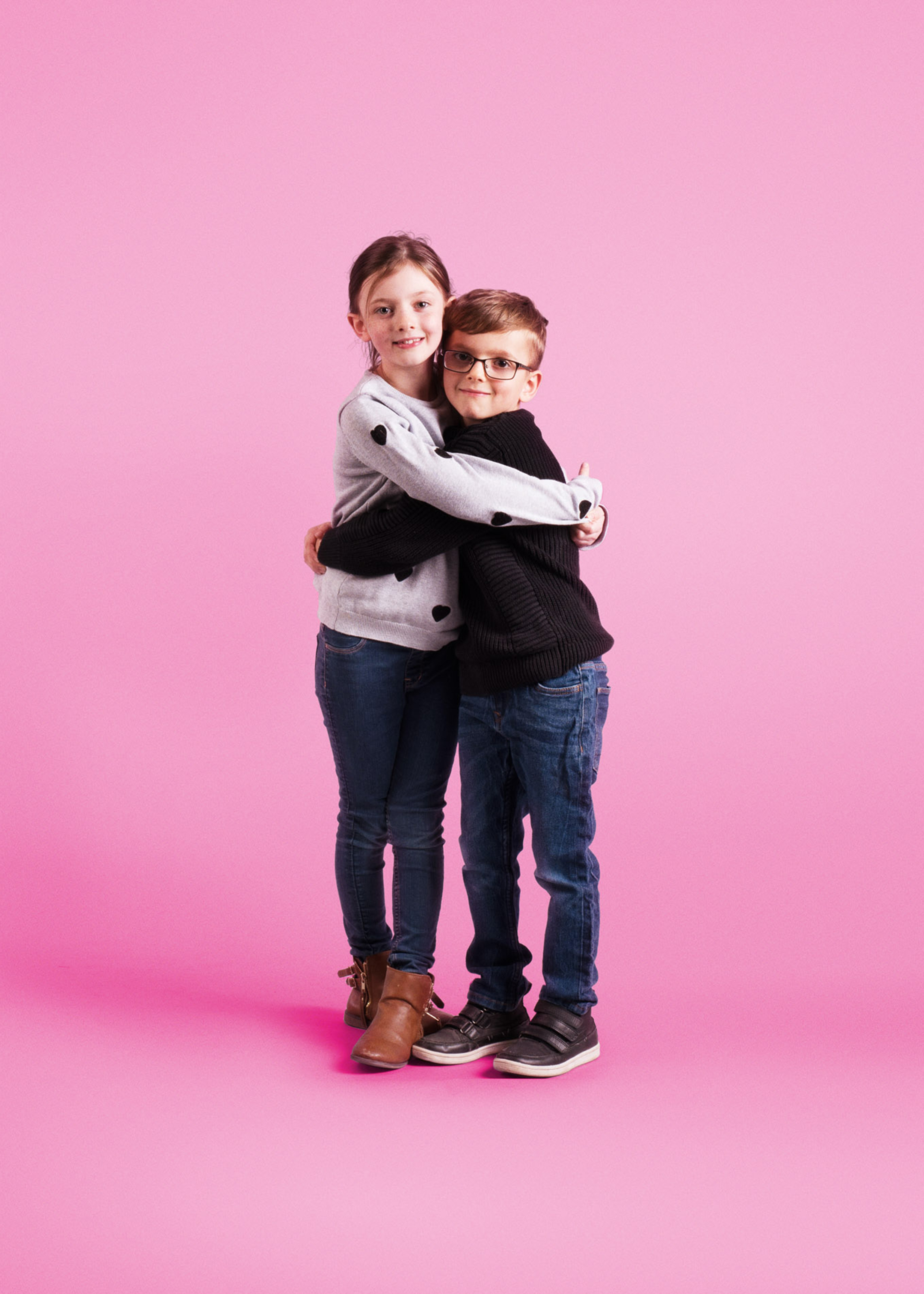 A young brother and sister hugging and smiling. They have been photographed on a pink background.