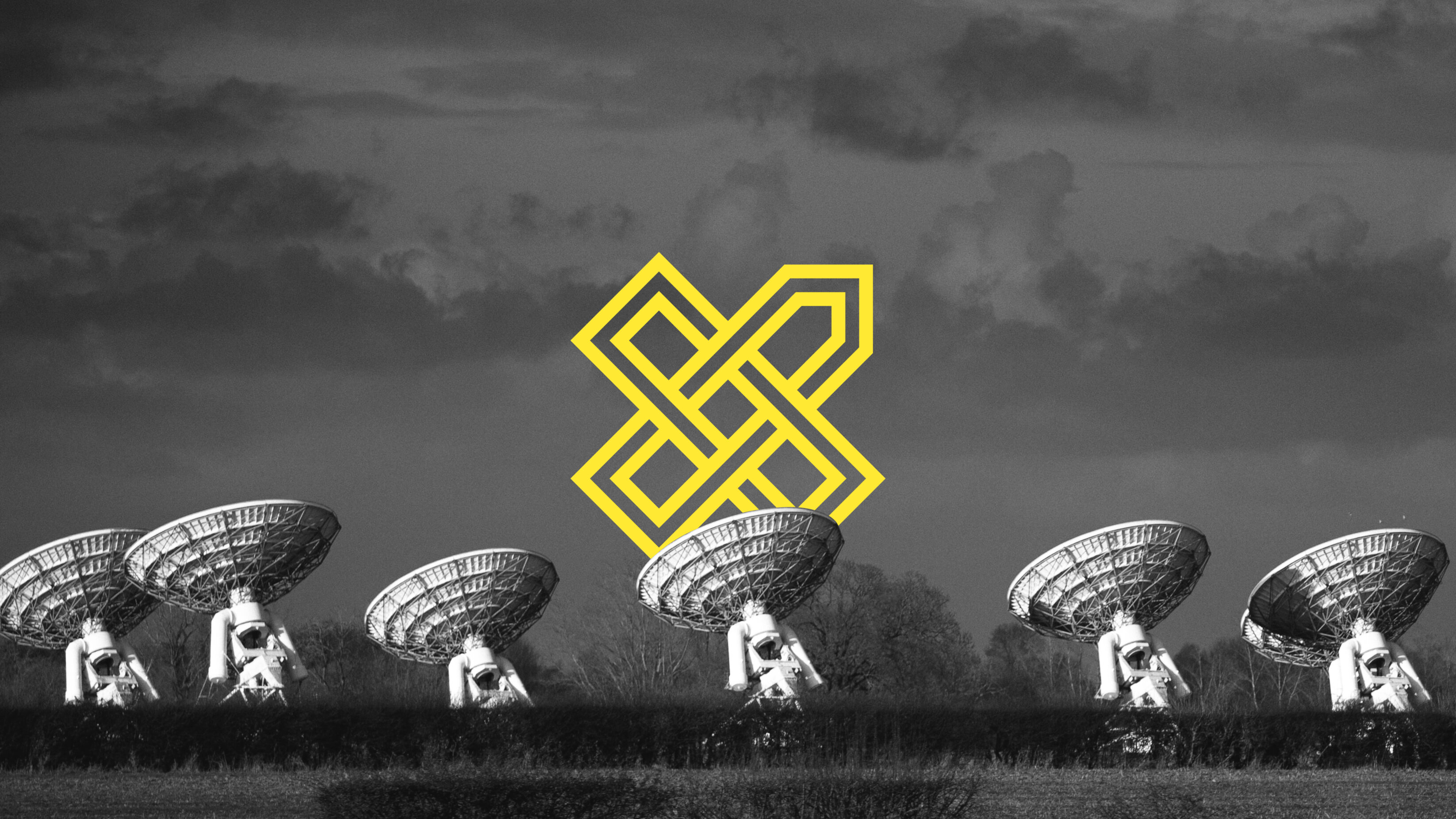 A series of satellite dishes facing away to a clouded sky. Image in black and white with the yellow Vox.Bio outline logo in the background.