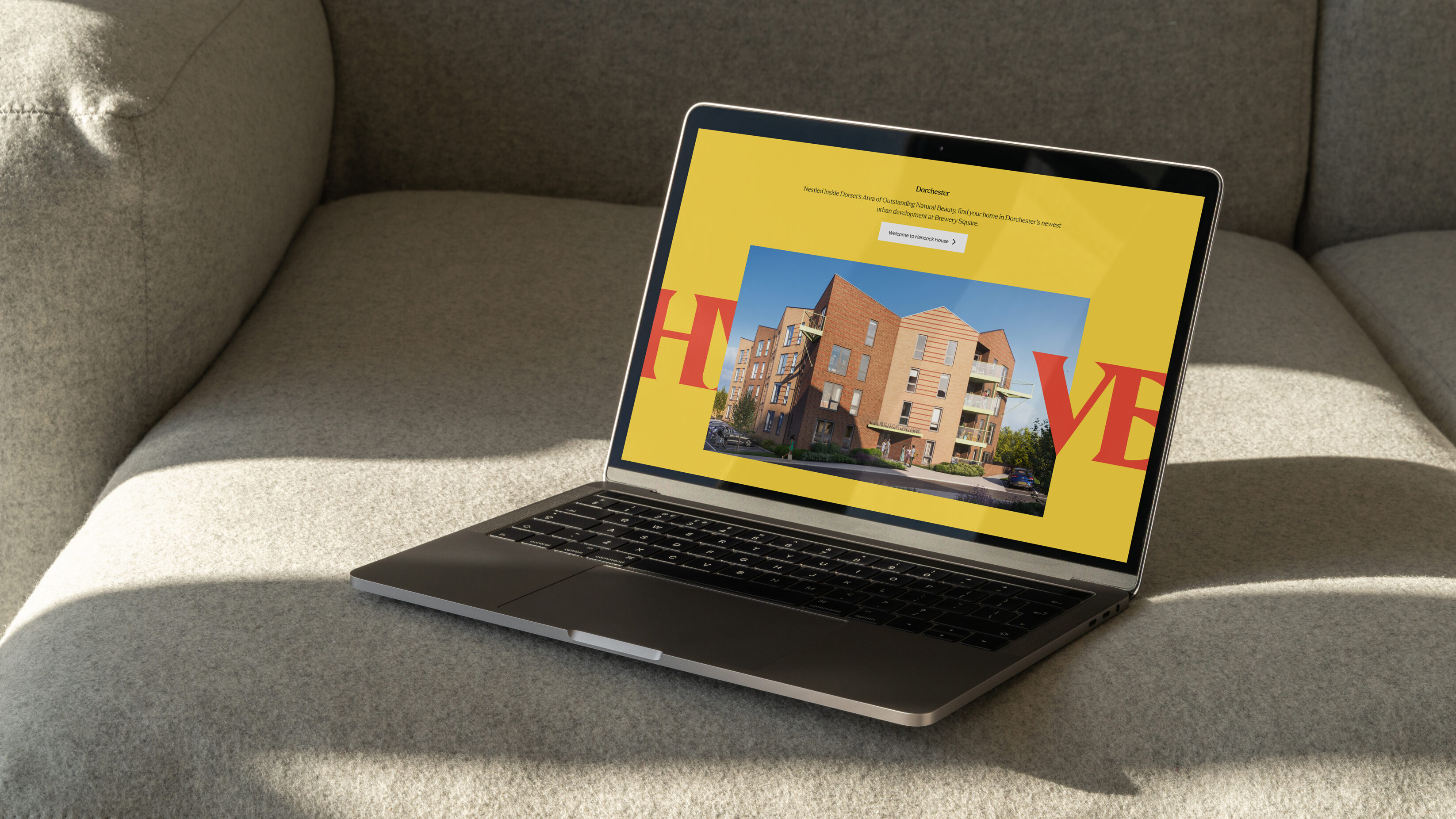 Website homepage visualised on a laptop, sitting on a light grey sofa, with shadows cast across the image.