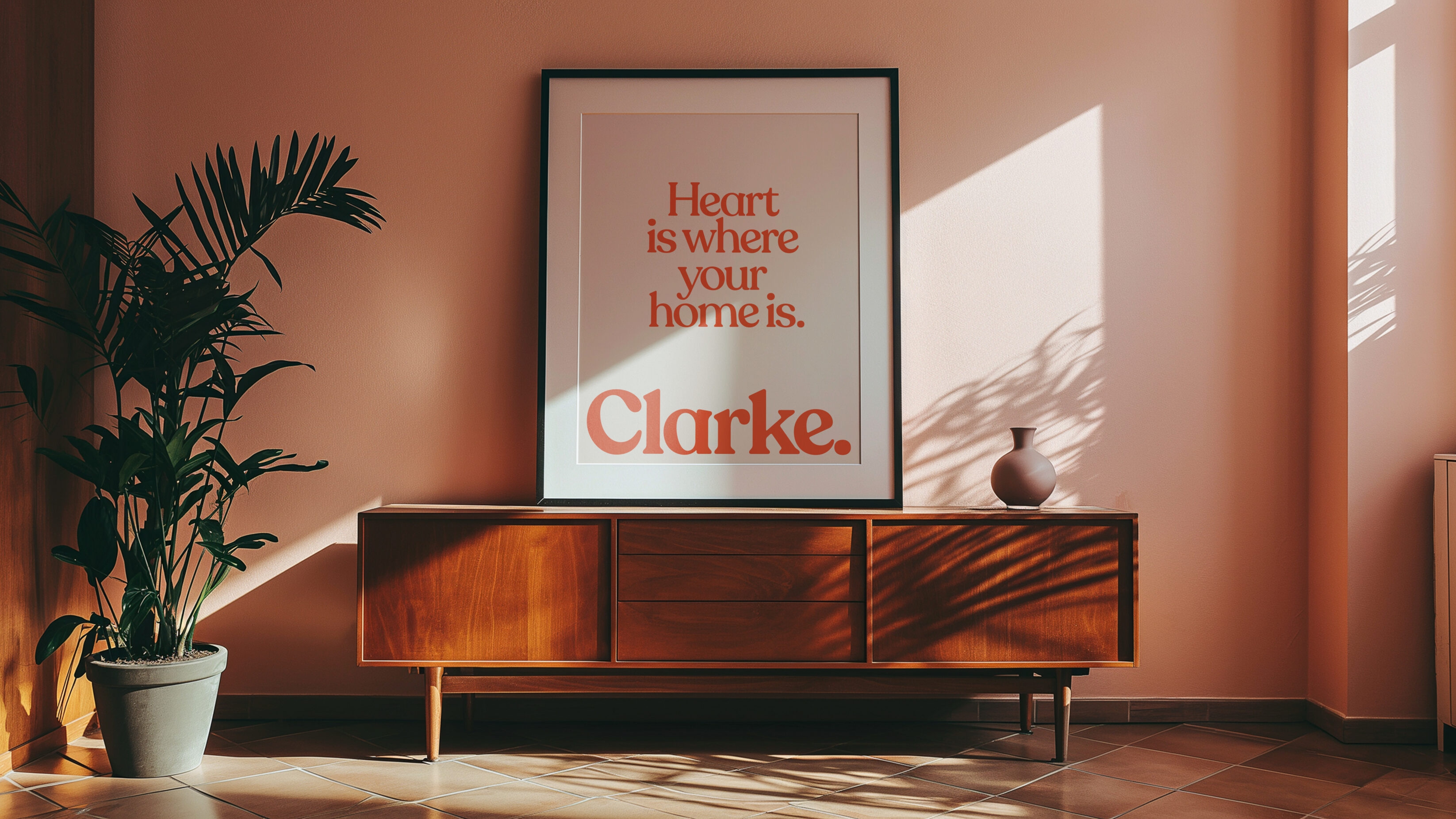 Framed poster in a black frame, on a dark wood cabinet, with the type 'Heart is where your home is.' written in a coral colour.