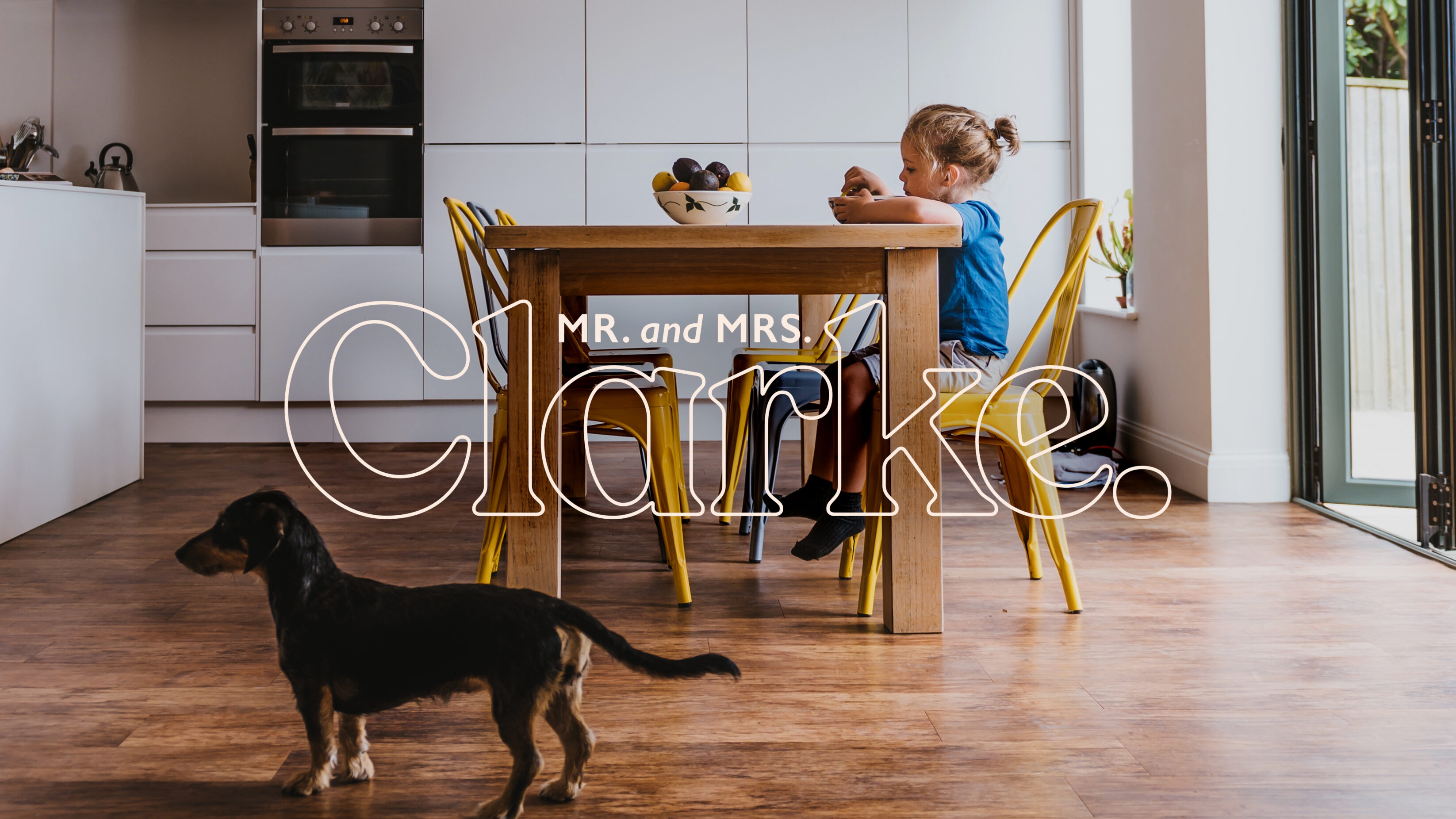 Kitchen photo, with a young girl sitting at a table and dog wandering around nearby. A large outline Mr and Mrs Clarke sits over the top. 