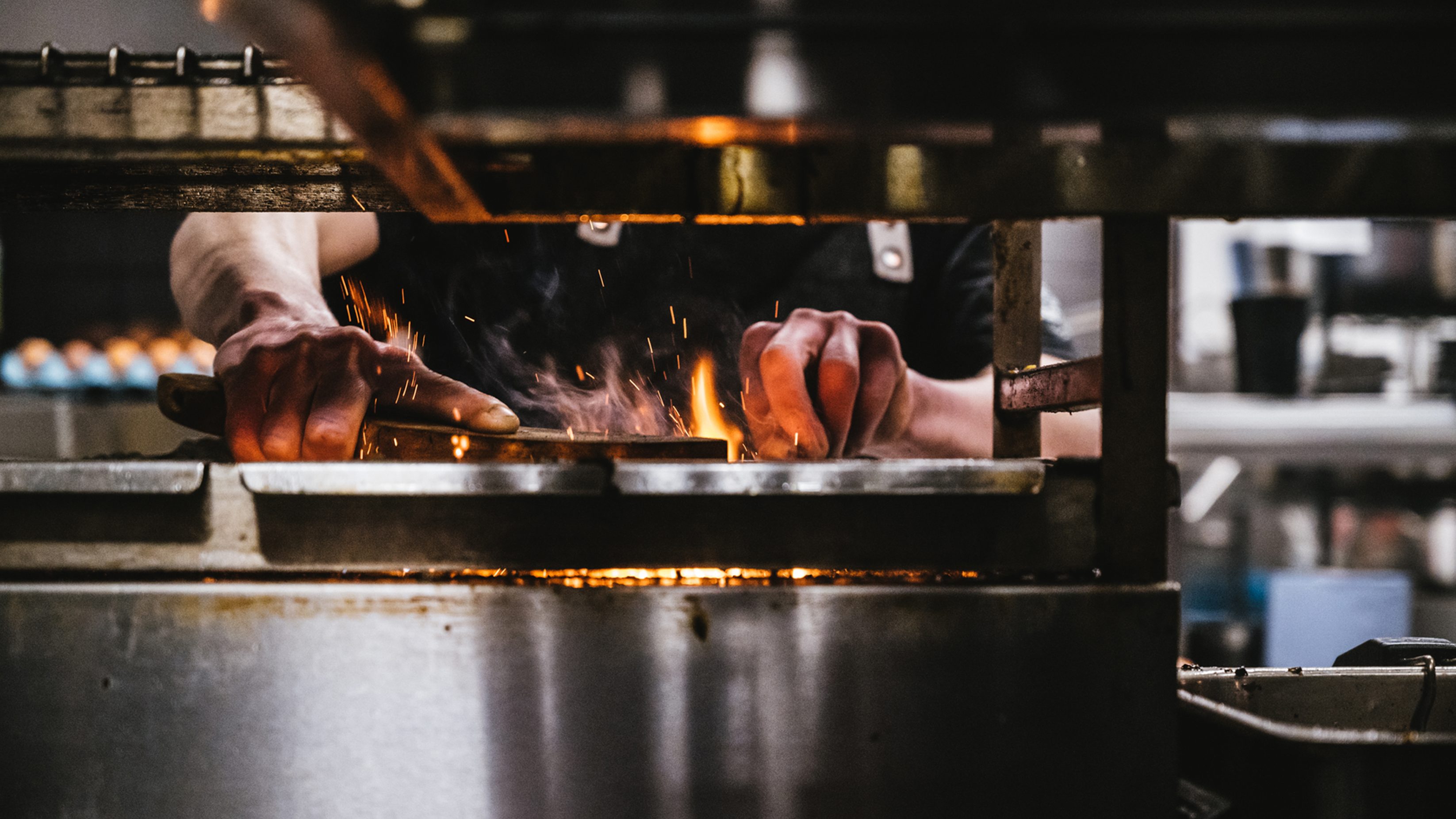 A chef's hands work with food over an open fire within the restaurants kitchen. The camera has caught this through the shiny metal shelving in the foreground.